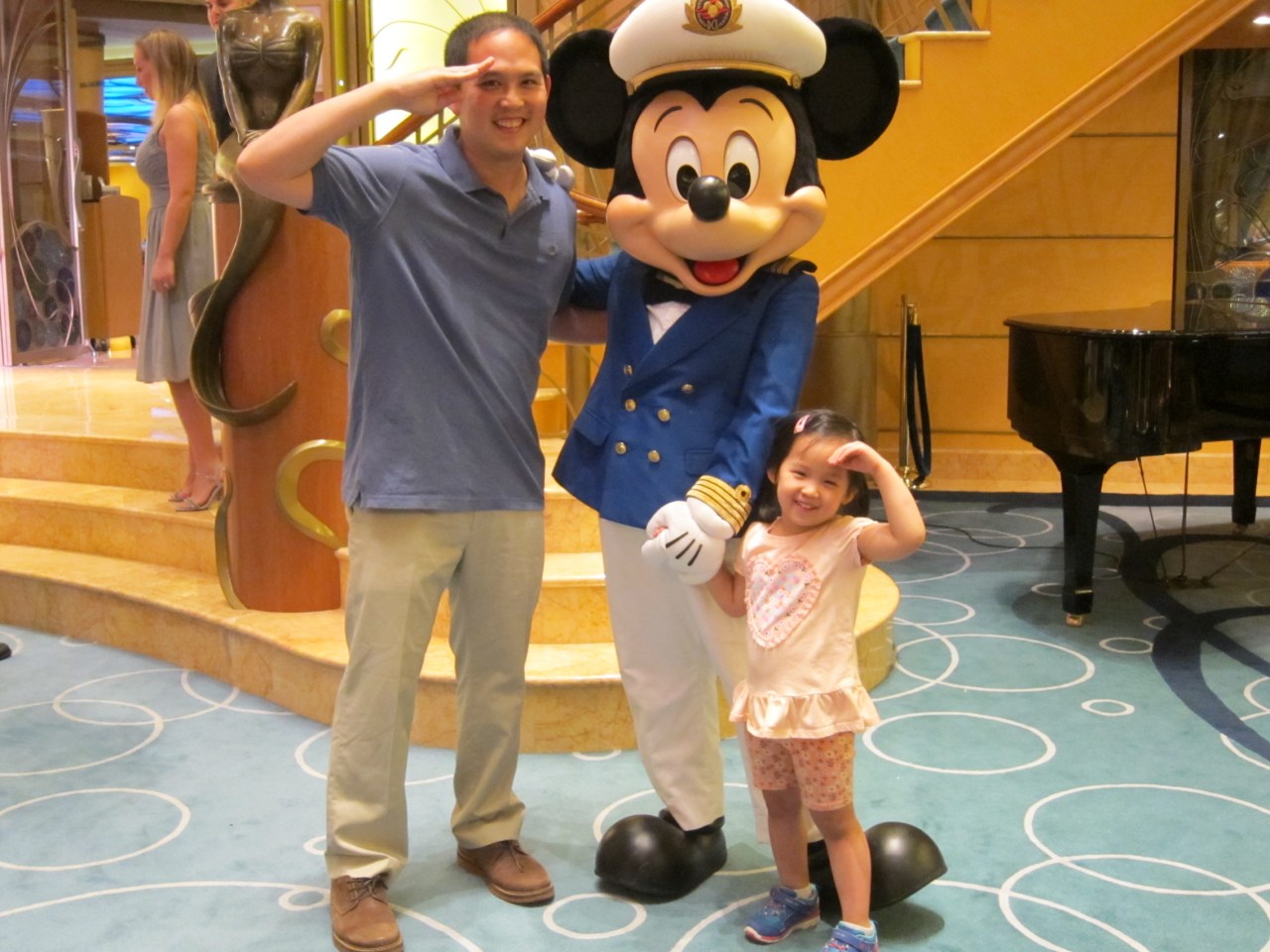 A man and a young girl are posing with a person dressed in a Mickey Mouse costume wearing a captain's uniform. They are all making a saluting gesture. The background includes a staircase, a piano, and a statue. The setting appears to be indoors, possibly in a themed venue or on a cruise ship.